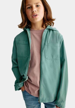 Chaqueta de cremallera verde claro con capucha, dos bolsillos en el pecho, utilizada sobre una camiseta rosa, combinada con jeans azules. Tejido suave y texturizado.