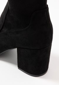 Gros plan sur une botte à talon carré en velours noir, montrant le talon et le matériau texturé sur fond blanc.