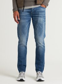 Vaqueros de mezclilla azules con un corte slim, que presentan áreas desgastadas y un sutil efecto de arrugas. Combinados con zapatillas grises y una camisa de manga larga gris.