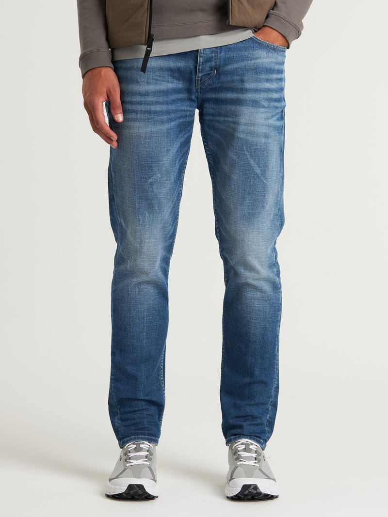 Vaqueros de mezclilla azules con un corte slim, que presentan áreas desgastadas y un sutil efecto de arrugas. Combinados con zapatillas grises y una camisa de manga larga gris.