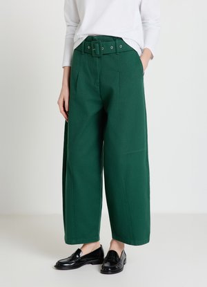Pantalon vert à jambes larges avec une taille ceinturée, présentant une texture lisse et de grandes poches latérales, associé à des mocassins noirs.