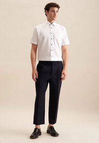 Wit shirt met korte mouwen en zwarte knopen, gecombineerd met marineblauwe taps toelopende broek en zwarte schoenen, tegen een neutrale achtergrond.