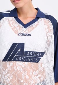 Balta mežģīņu blūze ar tumši zilu apkakli un piedurknēm, ar lielu tumši zilu logotipu "ADIDAS ORIGINALS" priekšpusē. Redzama sarežģīta mežģīņu raksts.