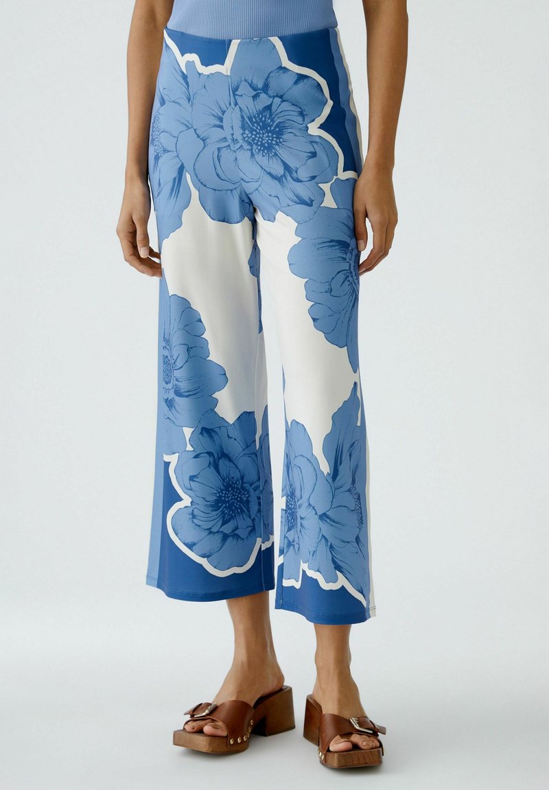 Weit geschnittene Hosen in Weiß und Blau mit großen Blumenmustern; glattes Material mit glänzender Oberfläche; getragen mit braunen Plateaustiefeln.