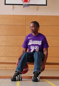 Violetiniai Lakers marškiniai su baltais ir geltonais akcentais, mėlynos džinsai ir juodi sportbačiai. Sėdi ant medinės suoliuko šalia krepšinio.
