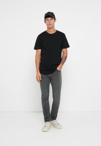 Hombre vestido con gorra negra, camiseta negra, jeans grises y zapatillas blancas, de pie contra un fondo blanco liso con una mano en el bolsillo.