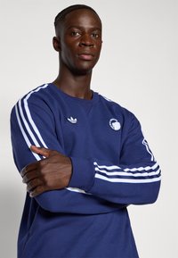 Jūrinis džemperis su baltais dryžiais, apvaliu kaklu ir logotipo pleistru. Pagamintas iš tekstūruoto audinio, su ilgomis rankovėmis ir gumbinėmis rankovėmis.