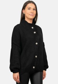 Zwarte chunky cardigan met hoge kraag, voorzien van grote zilverkleurige knopen, een geribbelde textuur en ballonsleeves.