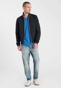 Veste légère noire avec col montant, doublure intérieure bleue, assortie à un t-shirt bleu et à un jean denim clair déchiré. Baskets blanches.