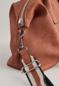 Braune Lederhandtasche mit strukturiertem Finish, ausgestattet mit einem silbernen Reißverschluss und einem abnehmbaren Tragegurt mit gestreiftem Muster in Creme und Marineblau.