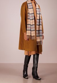 Mosterdkleurige wollen mantel, beige jurk met open hals, geruite sjaal met franjes, zwarte leren laarzen tot aan de knie met zilveren accenten.