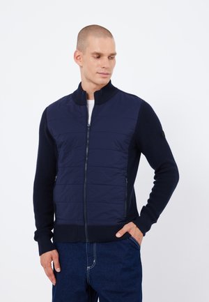 Navy jacket with front quilted panel and ribbed sleeves, features a high collar and zip closure. Two side pockets for storage.