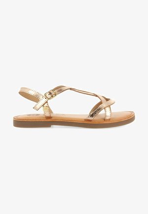 Sandalias de bronce metálico con múltiples correas cruzadas, hebilla ajustable en el tobillo y suela plana marrón. Textura de cuero suave.