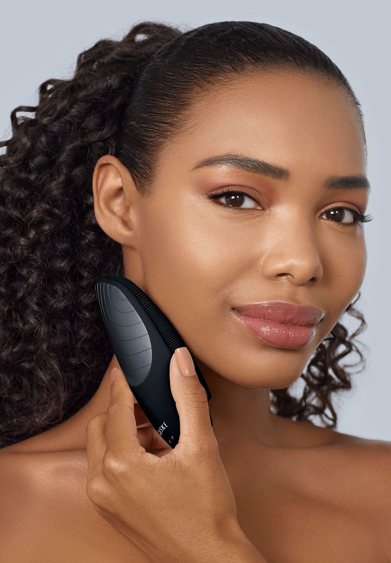 Woman with curly hair holds black facial cleansing brush against her right cheek, showing skincare routine.