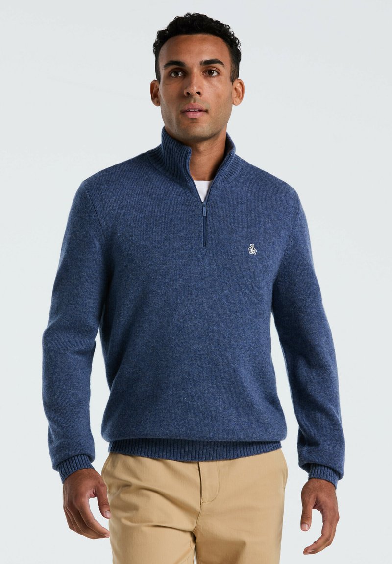 Man in een blauw sweater met kwartrits en een pinguïnlogo, met beige broek, staande tegen een effen lichte achtergrond.