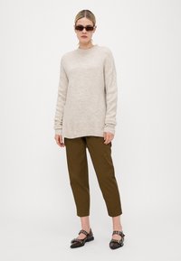 Beige strikket genser med ribbet kant, olivengrønne skreddersydde bukser og svarte sko med dekorative metall detaljer.