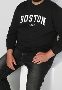 Černá mikina s kulatým výstřihem s nápisem "BOSTON PARK" v bílé barvě, spárovaná s tmavými džíny. Model sedí s rukama položenýma na stehně.