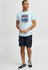 Világoskék póló, retro furgon grafikával piros és sötétkék színben. Sötétkék cargo sorttal párosítva, cipzáras zsebekkel és fekete sportcipővel.