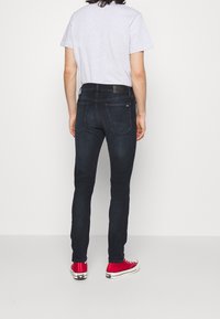 Vaqueros de denim oscuros con un corte ajustado, que presentan una cinturilla con un parche de cuero y bolsillos traseros. Combinados con zapatillas rojas y una camiseta gris.