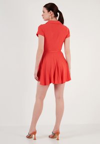 Red short-sleeve dress with a collared neckline, flared skirt, and textured fabric. Paired with orange heels and a colorful earring.