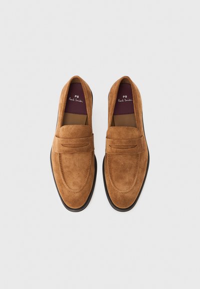 Smeđe mokasine od brušene kože s pravokutnim remenom preko gornjeg dijela, prošivenim detaljima i crnim gumenim potplatom. Unutrašnjost prikazuje uložak s brendom.