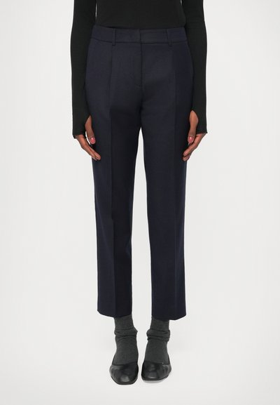 Navy tailored trousers with a straight cut, featuring front pleats and side pockets, made from a smooth fabric, paired with dark gray socks.