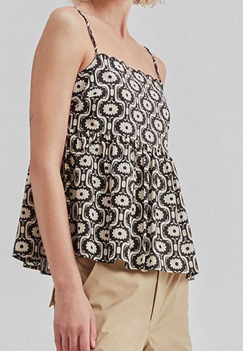 Femme portant un haut sans manches à motifs floraux noir et blanc avec un col en dentelle et un pantalon beige sur fond uni.