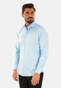 Camicia a maniche lunghe azzurro chiaro, con bottoni sul davanti, colletto classico e polsini a contrasto. Il tessuto appare liscio con una sottile trama.