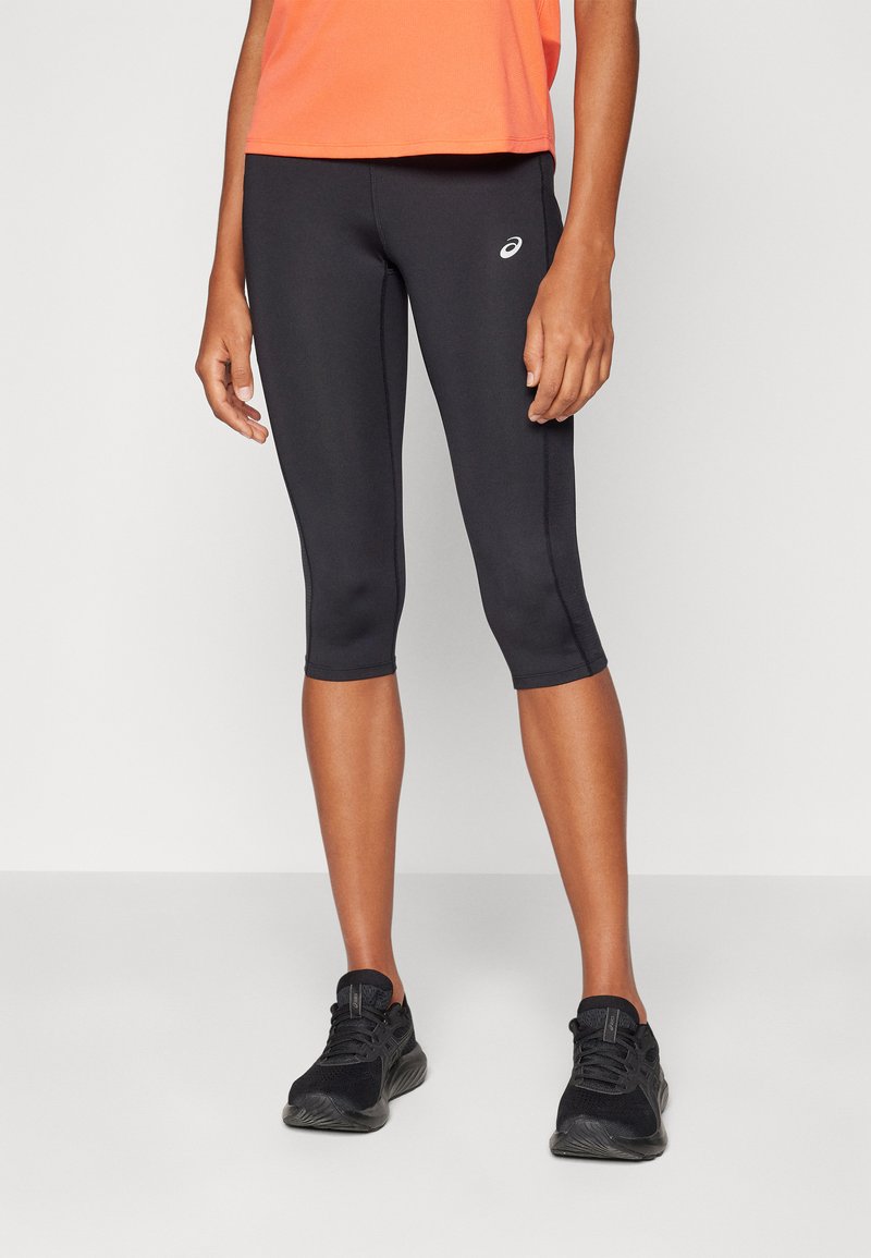 Leggings neri a vita alta realizzati in materiale elastico, caratterizzati da un design aderente e un sottile logo bianco su un lato, abbinati a sneakers nere.