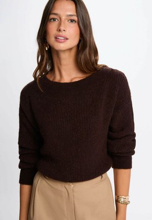 Maglione lavorato a maglia di colore marrone scuro con scollatura ampia e polsini arrotolati, abbinato a pantaloni beige a vita alta. Texture morbida, vestibilità casual.