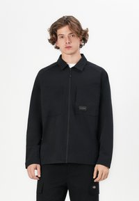 Chaqueta negra de cremallera hecha de una tela suave, con dos bolsillos en el pecho y un parche con logotipo. La chaqueta tiene un corte relajado y un cuello puntiagudo.