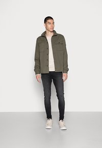 Camisa verde-oliva de botões com colarinho espalhado, duas Bolsos frontais, usada sobre uma t-shirt bege clara, combinada com jeans escuros e ténis claros.