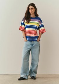 Modra majica s kratkimi rokavi z vodoravnimi rumenimi, rdečimi in belimi črtami. Oblečena z ohlapnimi svetlo modrimi kavbojkami.