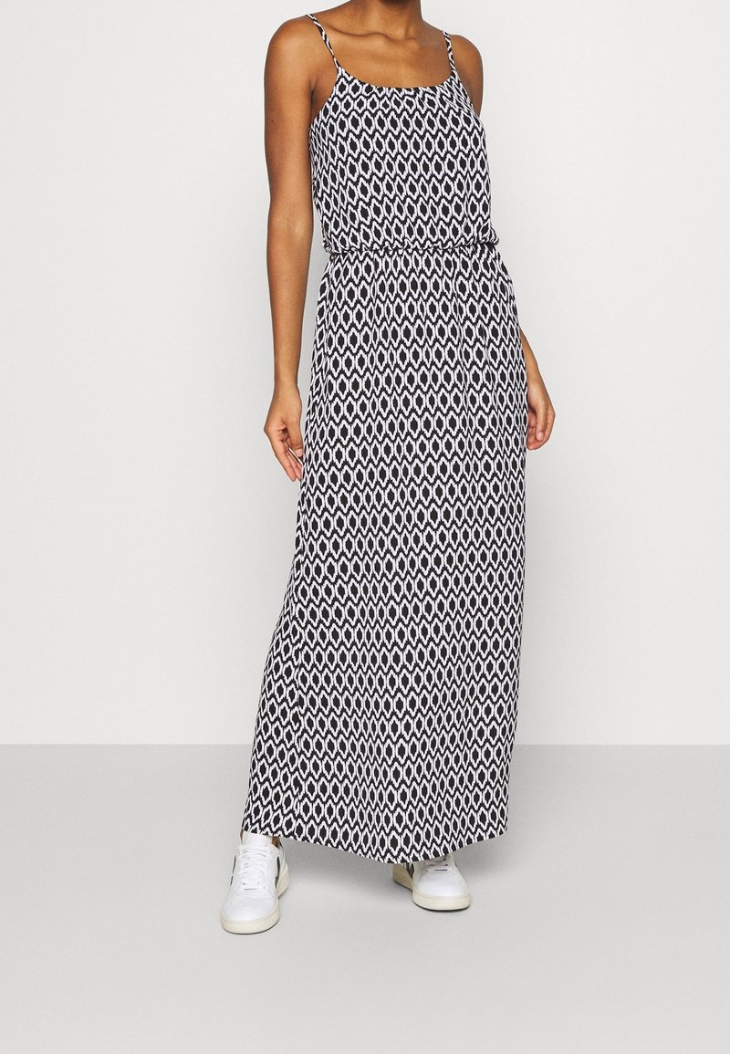 Robe maxi avec un motif géométrique noir et blanc, en tissu léger, bretelles spaghetti et taille élastique, associée à des baskets blanches.