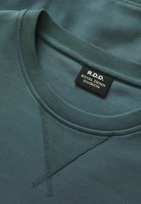 Türkisfarbenes Kurzarmshirt mit einem gerippten Rundhalsausschnitt, das ein gesticktes V-förmiges Design auf der Vorderseite und ein Etikett mit der Aufschrift "R.D.D. Royal Denim Division" aufweist.