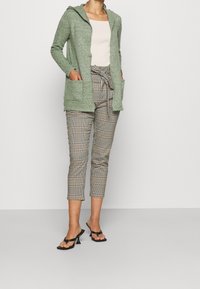 Světlý zelený kardigan s kapucí a dvěma předními kapsami, v kombinaci s vysokým pasem a károvanými kalhotami, a černými sandály na podpatku.