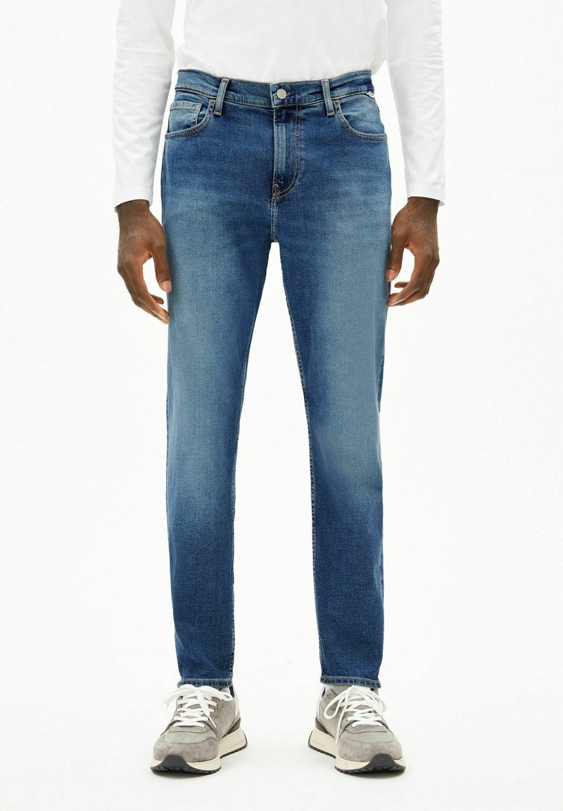 Homme portant un jean slim bleu moyen, une chemise blanche à manches longues et des baskets grises, debout face à l'avant sur un fond blanc.
