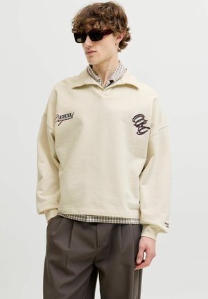 Giovane uomo con capelli ricci che indossa occhiali da sole tartarugati, una felpa beige sopra una camicia a quadri e pantaloni scuri plissettati, in piedi con le mani nelle tasche.