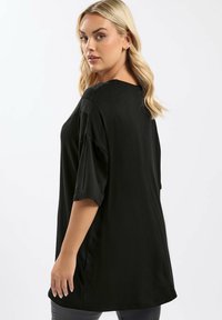 Kvinna med blont hår som bär en lös svart t-shirt, vänd något på sidan, och tittar över sin axel mot en enfärgad vit bakgrund.