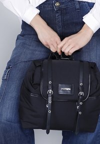 Mochila negra de nylon con acentos de cuero, que presenta una solapa frontal, dos correas y una placa de marca. Asa de transporte en la parte superior.