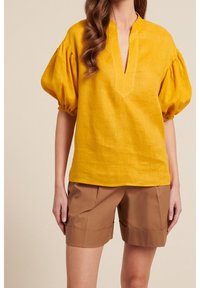 Blusa in lino giallo senape con maniche a sbuffo, scollatura a V e orlo dritto, abbinata a pantaloni corti marroni su misura. Tessuto morbido, design minimalista.