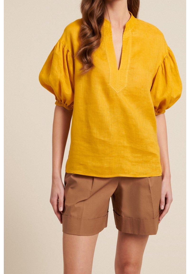 Blusa in lino giallo senape con maniche a sbuffo, scollatura a V e orlo dritto, abbinata a pantaloni corti marroni su misura. Tessuto morbido, design minimalista.