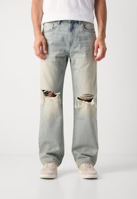 Jeans de mezclilla azul claro con un corte relajado, detalles desgastados que incluyen grandes desgastes en las rodillas y bordes deshilachados. Llevados con zapatillas beige.
