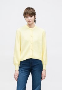 Κίτρινη ζακέτα με στρογγυλή λαιμόκοψη, υφή με ριγέ μοτίβο και κουμπιά με πέρλες, συνδυασμένη με μπλε τζιν παντελόνι.