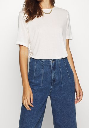 Biała koszulka z krótkim rękawem w połączeniu z ciemnoniebieskimi dżinsami z wysokim stanem. Koszulka ma prosty, gładki wzór; dżinsy posiadają przednie zakładki.