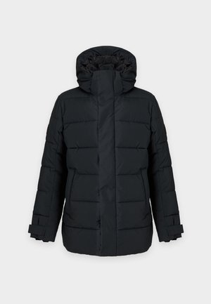 Homme debout portant une veste matelassée noire, une chemise beige, un pantalon foncé, des chaussettes blanches et des baskets noires sur un fond uni.