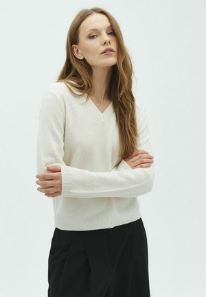 Femme aux longs cheveux châtain clair portant un pull blanc à col en V et un pantalon noir, debout les bras croisés devant un fond uni.
