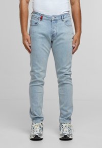 Férfi világoskék szűk farmert, fehér trikót és fehér sportcipőt visel, kék és szürke részletekkel, szürke háttér előtt áll.