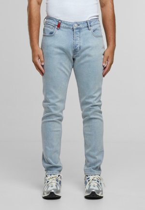 Mies, joka on pukeutunut vaaleansinisiin slim fit -farkkuihin, valkoiseen tanktoppiin ja valkoisiin lenkkareihin, joissa on sinisiä ja harmaita yksityiskohtia, seisoo harmaata taustaa vasten.