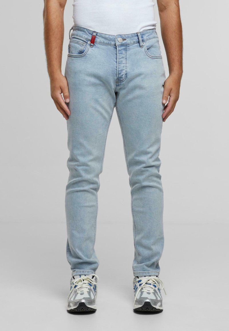 Férfi világoskék szűk farmert, fehér trikót és fehér sportcipőt visel, kék és szürke részletekkel, szürke háttér előtt áll.